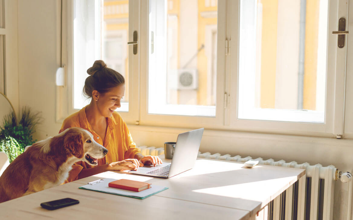 A woman with a dog beside her is sitting at a table and looking at a laptop. 