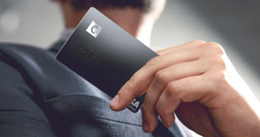 A man in a suit is holding a black Citadele C Infinite payment card with the C logo in the top left corner, focusing his attention on the card.