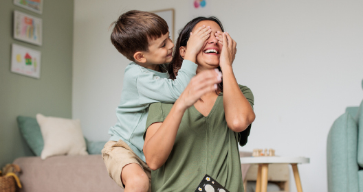Šeima sėdi prie stalo. Mama su kūdikiu rankose geria kavą ir žiūri į nešiojamąjį kompiuterį, o tėtis moko kitą vaiką piešti. 