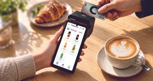 A customer taps a Citadele payment card on a payment terminal in a café, with a cappuccino and a croissant on the table.