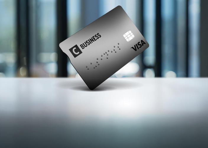 Pilka C Business kreditinė kortelė, pastatyta vertikaliai ant paviršiaus, modernaus biuro fone.