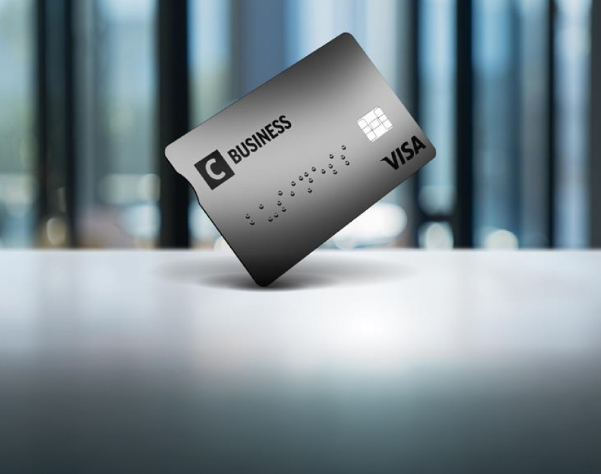 Pilka C Business kreditinė kortelė, pastatyta vertikaliai ant paviršiaus, modernaus biuro fone.