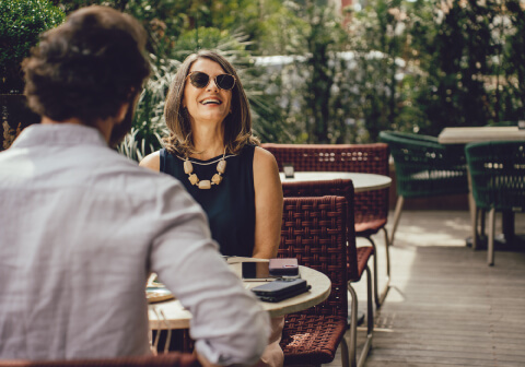 A woman in sunglasses sits at an outdoor café table, smiling and talking to a man seen from behind.