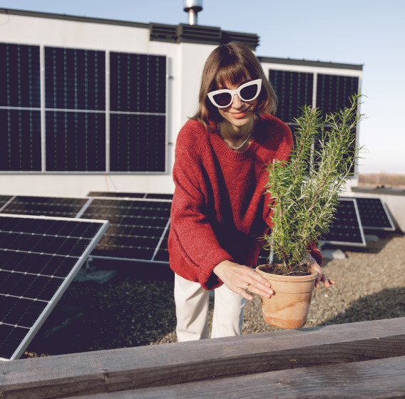 A girl wearing sunglasses is holding a green plant while standing on a rooftop terrace of a house equipped with solar panels. 