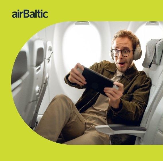 Žaliame fone airbaltic logotipas ir nuotrauka, kurioje vyras sėdi lėktuve ir žaidžia su planšete.