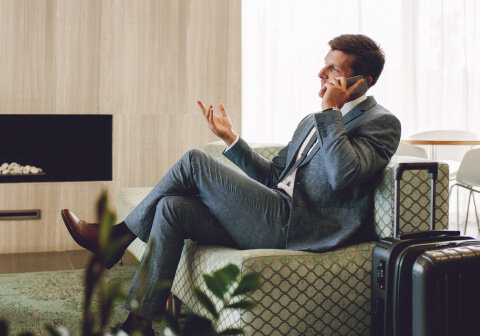Official Priority Pass image – a person is seated in a Priority Pass lounge, speaking on the phone; a travel suitcase is placed beside them.