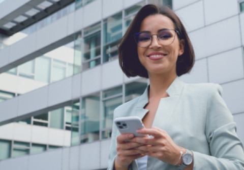 A woman in business attire is standing by a modern glass building, holding a smartphone.