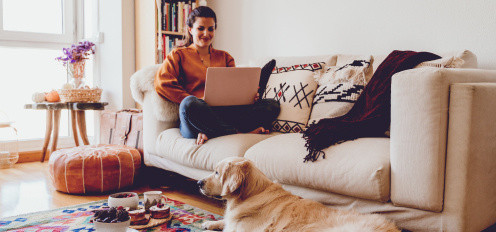 Moteris sėdi ant šviesios sofos su nešiojamuoju kompiuteriu, o ant spalvingo kilimo priešais guli auksaspalvis retriveris jaukiame, šviesiame svetainės kambaryje.